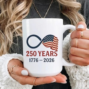 May include: White ceramic mug featuring the text "250 YEARS 1776-2026" with an infinity symbol and a waving American flag design. The mug has a large handle and is held by a person.