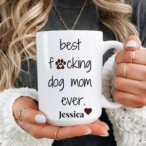 May include: White ceramic mug with the text "best f**king dog mom ever." The word "f**king" has a paw print in place of the "o". The name "Jessica" is below the text, with a heart symbol.