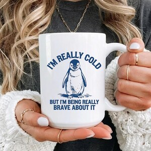 May include: White ceramic mug with a blue penguin illustration and the text "I'M REALLY COLD BUT I'M BEING REALLY BRAVE ABOUT IT". The mug has a large handle and is held by a person.