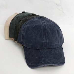 May include: A stack of baseball caps in various colors: navy blue, dark green, charcoal, and tan. The caps have a classic, unstructured design with a curved brim. The fabric appears to be a washed cotton or similar material, giving them a vintage look.