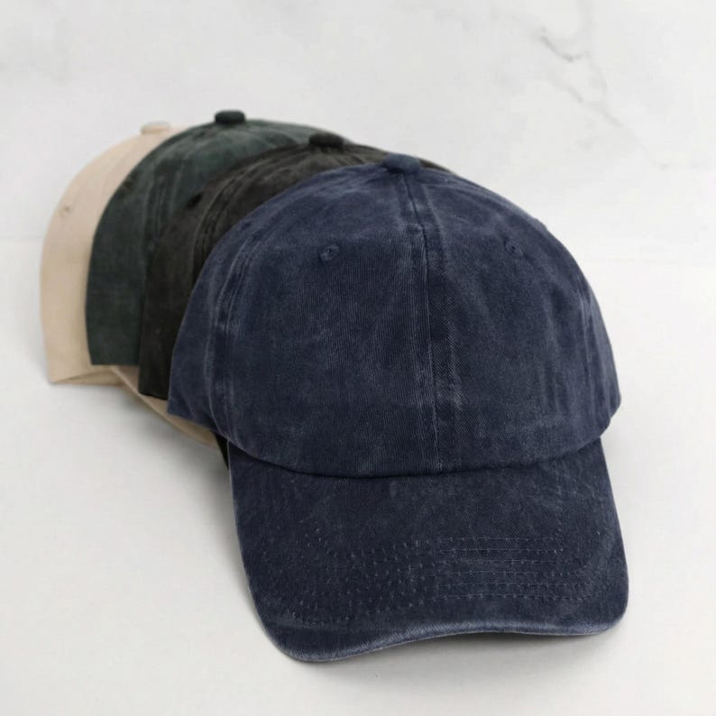 May include: A stack of baseball caps in various colors: navy blue, dark green, charcoal, and tan. The caps have a classic, unstructured design with a curved brim. The fabric appears to be a washed cotton or similar material, giving them a vintage look.