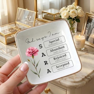 May include: A white ceramic trinket dish with gold trim, featuring the text "God says I am" and the name "SARA" with inspirational words. A pink flower and leaves are painted on the dish. The dish is held by a person.