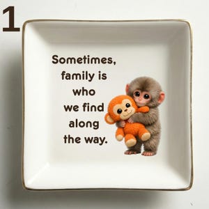 May include: Square ceramic trinket dish with a gold rim. The dish features a graphic of two cartoon monkeys hugging, with the text "Sometimes, family is who we find along the way."