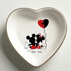 May include: Heart-shaped white ceramic dish with a gold rim. Features a black and red cartoon couple embracing, holding a heart-shaped balloon. The names "Noah Sarah" are printed below the couple.
