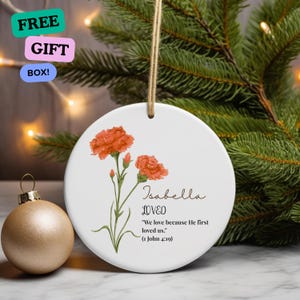 May include: White ceramic Christmas ornament with a floral design and the name "Isabella". The ornament features orange flowers and the text "LOVED" and a Bible verse. A gold ornament is to the left.
