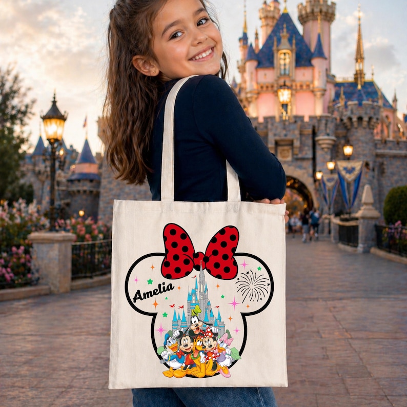 May include: A cream-colored tote bag featuring a Minnie Mouse design. The design includes a red polka-dot bow, a castle, and various Disney characters. The name "Amelia" is printed on the bag. The bag has two white straps.