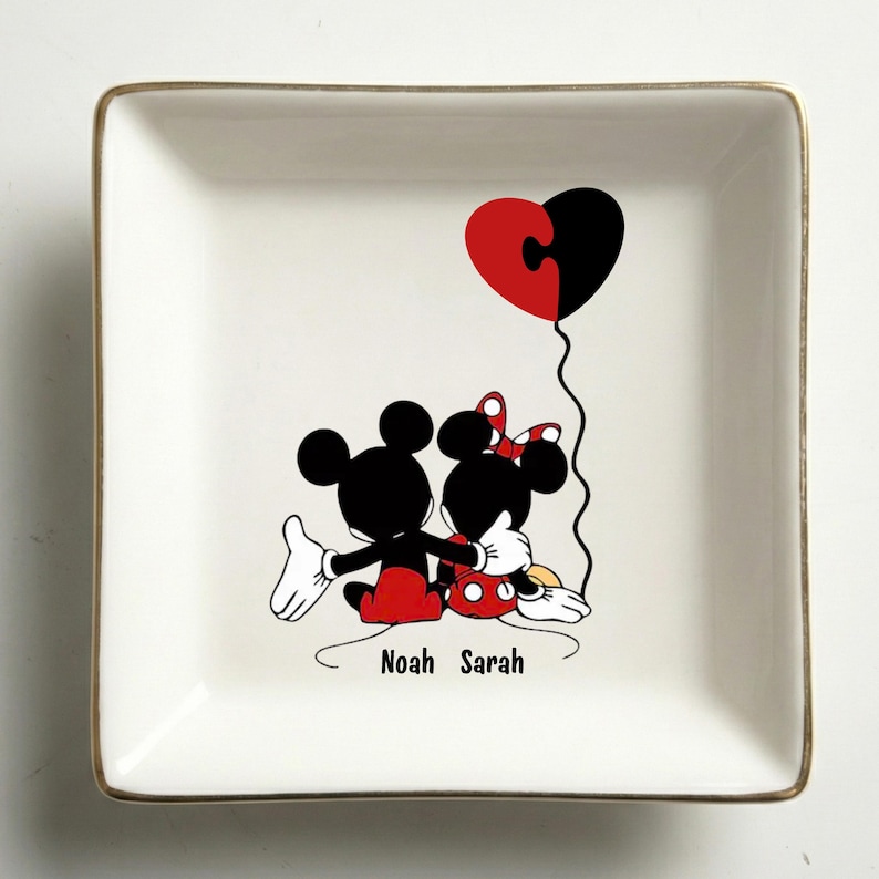 May include: Square white ceramic dish with a gold rim, featuring a cartoon illustration of Mickey and Minnie Mouse embracing. A heart-shaped balloon with puzzle piece design floats above them. The names "Noah Sarah" are printed below.