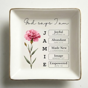 May include: A square, white ceramic dish with a gold rim. The dish features a pink carnation illustration and the words "God says I am." The name "JAMIE" is spelled out with words like "Joyful" and "Empowered."
