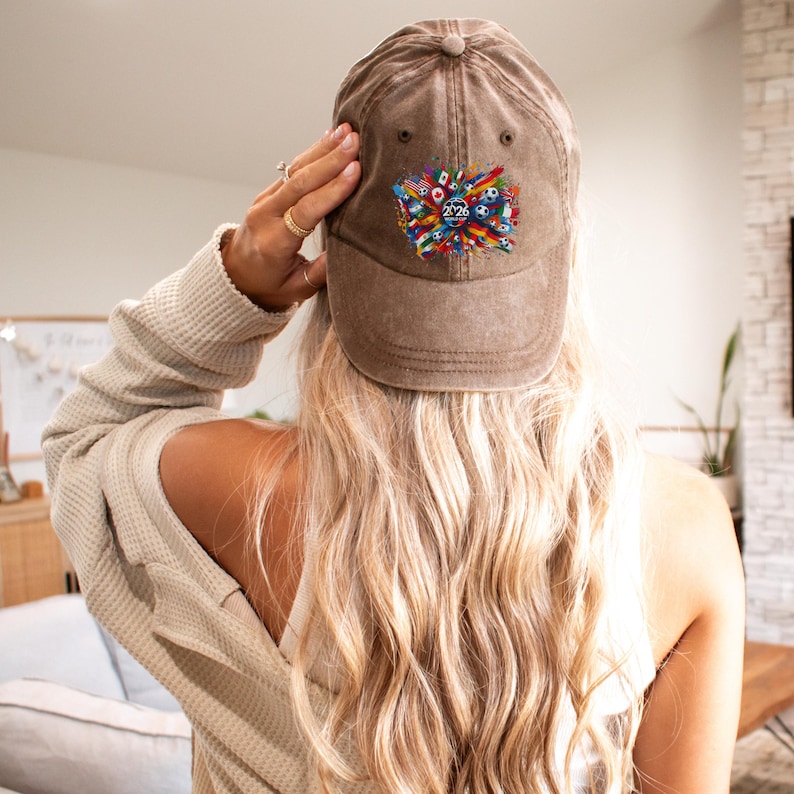 May include: A brown baseball cap with a colorful graphic design featuring flags, soccer balls, and the number "2026". The cap has a curved brim and a button on top. The cap is worn by a person.