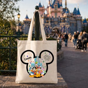 May include: Cream-colored tote bag featuring a black Mickey Mouse head design with the name "Liam" and a colorful illustration of Disney characters and a castle. The bag has a handle and is set against a blurred background.
