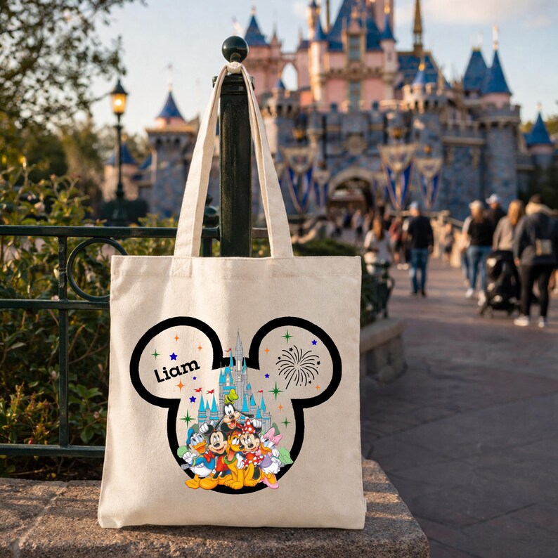 May include: Cream-colored tote bag featuring a black Mickey Mouse head design with the name "Liam" and a colorful illustration of Disney characters and a castle. The bag has a handle and is set against a blurred background.