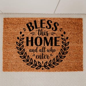 May include: A rectangular doormat with a brown coir surface. The mat features the words "BLESS this HOME and all who enter" in black, surrounded by a black laurel wreath design. The mat is placed on a light-colored, textured surface.