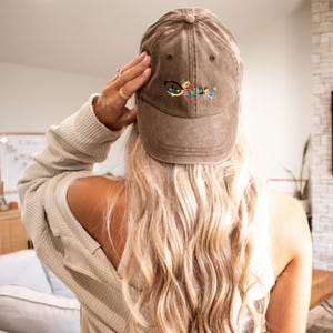 Op de afbeelding: Een bruine baseballcap met het woord "Disney" geborduurd in kleurrijke letters. De pet wordt gedragen door een persoon met lang blond haar, die een beige wafel gebreide lange mouwen shirt draagt.