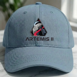 May include: A light blue baseball cap featuring the "Artemis II" mission logo. The logo is a black triangle with a moon and Earth design, red accents, and the names of the crew. The text "ARTEMIS II" is printed below the logo.