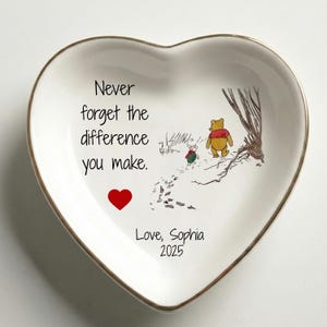 May include: Heart-shaped ceramic trinket dish with a gold rim. The dish features a Winnie the Pooh illustration with the text "Never forget the difference you make." A red heart and the text "Love, Sophia 2025" are also included.