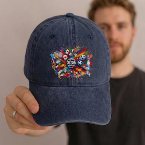 May include: A navy blue baseball cap featuring a colorful graphic design. The design includes flags, soccer balls, and the text "2026 WORLD CUP". The cap is made of a durable fabric and has a curved brim.