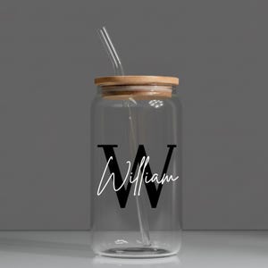 May include: A clear glass can-shaped tumbler with a wooden lid and a clear straw. The tumbler features a large black letter "W" with the name "William" in white script. The design is centered on the glass.