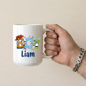 May include: A white ceramic mug with a black handle and the text "But First, Coffee" in a black, handwritten-style font. The mug is on a white surface.