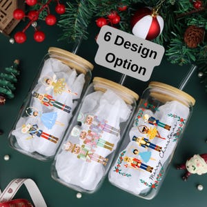 May include: Three glass tumblers with bamboo lids and straws, each decorated with a different Nutcracker-themed design. The tumblers are filled with white tissue paper. The text "6 Design Option" is in the center of the image. The background is a dark green.