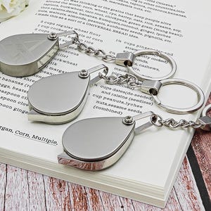 May include: Three silver-toned magnifying glass keychains with a teardrop-shaped design are displayed on a book. Each keychain has a chain and a large ring. The book's pages are open, and text is visible.
