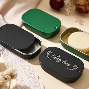 Peut inclure: Plusieurs boîtes ovales en métal noir et vert. L'une des boîtes noires est ouverte, révélant un intérieur argenté. Une autre boîte noire est gravée du nom "Angelina" et de motifs floraux. Un contenant vert contient un savon rond de couleur claire.