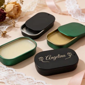 Pode incluir: Quatro latas ovais, duas pretas e duas verdes, com tampas. Uma lata preta tem o nome "Angelina" gravado. As latas contêm um bálsamo ou perfume sólido. Flores secas e fitas no fundo.