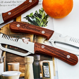 May include: A collection of kitchen tools with wooden handles and stainless steel blades. The tools include a can opener, a peeler, and a knife. An orange sits nearby. The word "Carolina" is engraved on one of the handles.