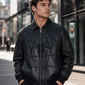 May include: A black leather bomber jacket featuring a prominent embossed spiderweb pattern across the front and sleeves, a classic collar, a full-length silver-toned metal zipper closure, and ribbed cuffs at the wrists and waist hem.