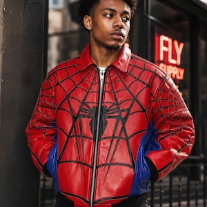 May include: A red and blue leather bomber jacket featuring a black spider web pattern across the chest and sleeves, a central black spider emblem, a silver front zipper, a classic collar, and black ribbed cuffs and hem at the waistline.