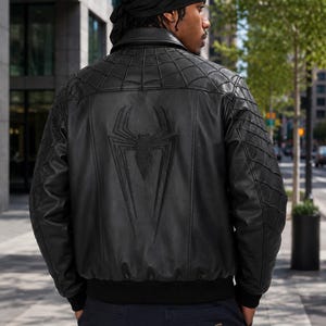 May include: A black leather bomber jacket featuring a large embossed spider emblem on the back. The upper back and sleeves have a stitched spiderweb pattern. The jacket includes a ribbed black waistband and cuffs, a classic collar, and a smooth finish.