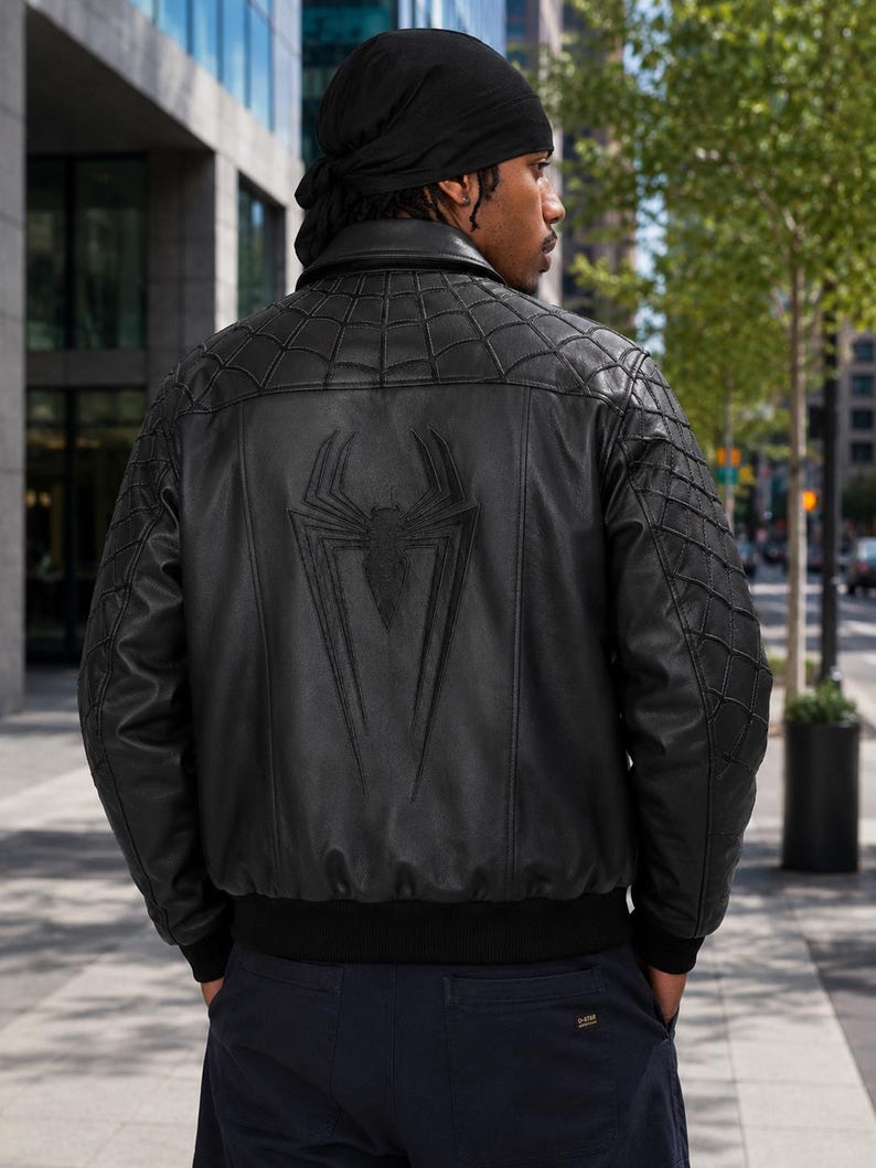 May include: A black leather bomber jacket featuring a large embossed spider emblem on the back. The upper back and sleeves have a stitched spiderweb pattern. The jacket includes a ribbed black waistband and cuffs, a classic collar, and a smooth finish.