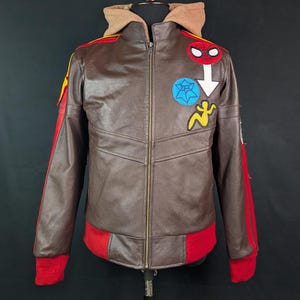 May include: A brown leather jacket featuring a tan hood, a front zipper, and red ribbed cuffs and hem. The chest displays embroidered patches including a red spider mask, a blue spiderweb circle, a white arrow, and a yellow stylized figure.