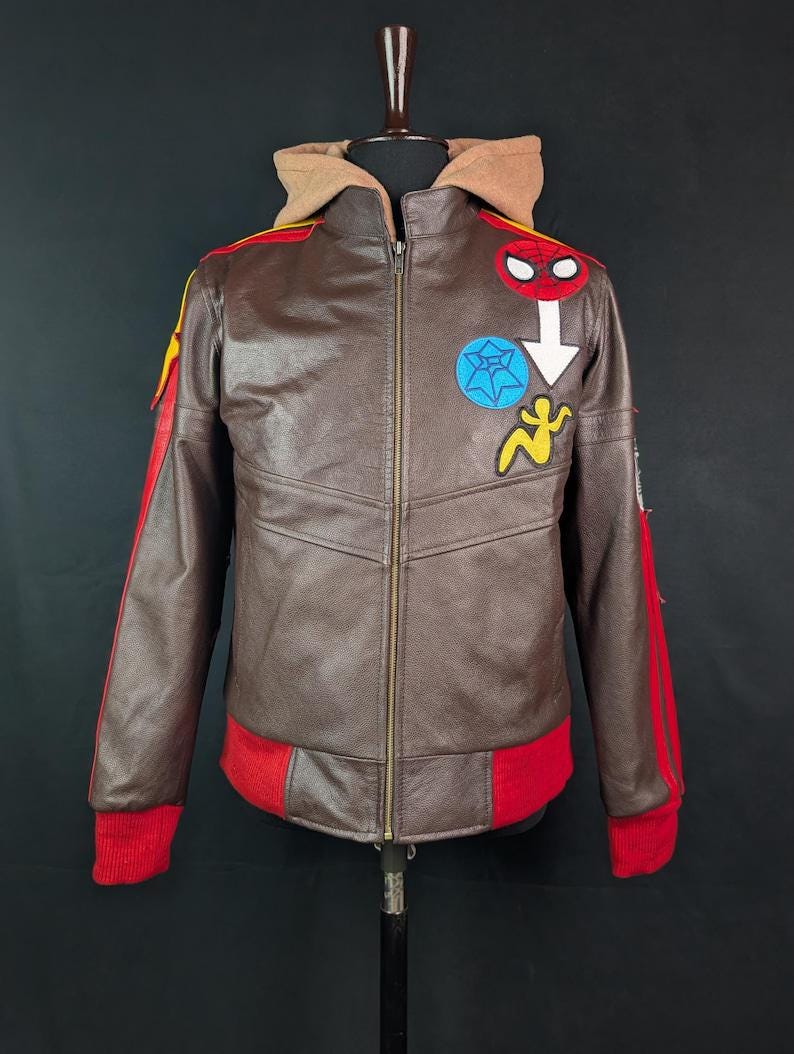 May include: A brown leather jacket featuring a tan hood, a front zipper, and red ribbed cuffs and hem. The chest displays embroidered patches including a red spider mask, a blue spiderweb circle, a white arrow, and a yellow stylized figure.