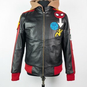 May include: A black leather jacket featuring a tan hood, a front zipper, and red ribbed cuffs and hem. The chest displays a red Spider-Man mask patch, a blue spiderweb patch, and a yellow figure patch. Red stripes run down the sleeves.