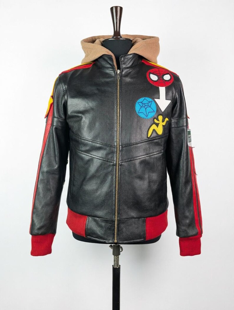 May include: A black leather jacket featuring a tan hood, a front zipper, and red ribbed cuffs and hem. The chest displays a red Spider-Man mask patch, a blue spiderweb patch, and a yellow figure patch. Red stripes run down the sleeves.