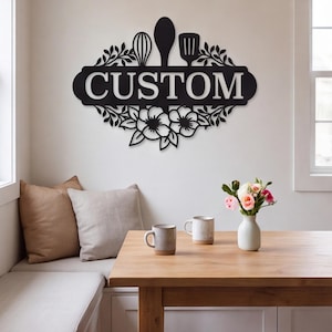 May include: A black metal wall sign featuring the word CUSTOM in bold capital letters. The design includes silhouettes of a whisk, a spoon, and a spatula at the top, with decorative leaf and flower motifs framing the text on the sides and bottom.