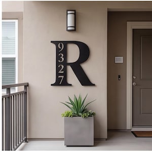 May include: A large, matte black metal wall sign featuring the capital letter R with the numbers 9, 3, 2, and 7 vertically aligned within the left side of the letter. The sign is mounted on a beige exterior wall above a square gray planter.