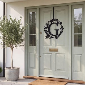 May include: A black metal door hanger featuring a circular wreath design with leaves surrounding a large capital letter G. The sign is suspended by a thin black vertical strap from the top of a light sage green paneled front door.