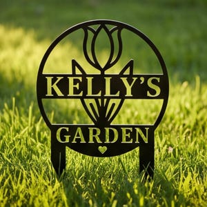 May include: A circular black metal garden sign featuring a cutout tulip design at the top. The text KELLY'S GARDEN is centred in the middle, with a small heart shape positioned below the word GARDEN. The sign has two stakes at the bottom.