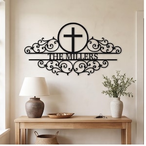 May include: A black metal wall sign featuring a central circle with a cross inside, flanked by symmetrical scrollwork patterns. The text THE MILLERS is centered horizontally across the design, integrated into the decorative metal structure.