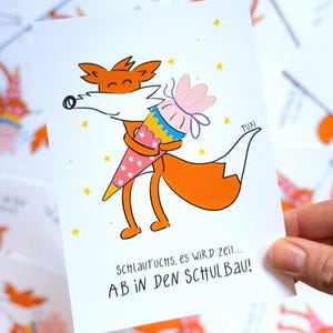 Könnte beinhalten: Eine weiße Karte mit einem orangefarbenen Cartoon-Fuchs, der eine rosa und blaue Schultüte hält. Der Fuchs hat eine weiße Brust und eine schwarze Nase. Der Text auf der Karte lautet "SCHLAUFUCHS, es wird Zeit... AB IN DEN SCHULBAU!"