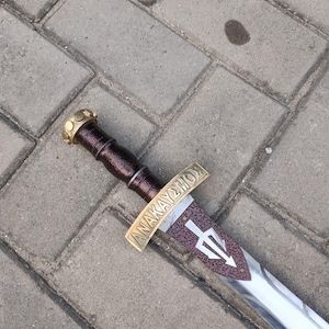 May include: A decorative sword with a brown handle and a gold-colored hilt. The blade is silver with a trident symbol and the text "ANAKAYΣMOΣ" on the hilt. The sword is lying on a gray brick surface.