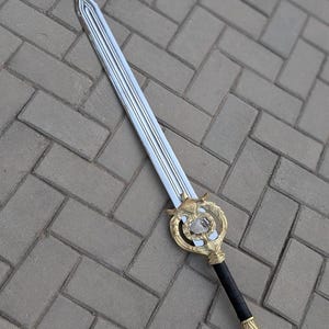 Puede incluir: Espada de fantasía plateada y dorada con una guarda decorativa con una gema transparente. La hoja tiene un canal central y el mango está envuelto en cuero negro. Fondo de ladrillos grises.