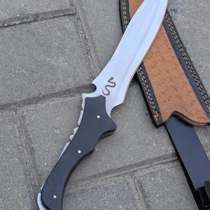 May include: A handmade knife with a silver blade and a black handle. The blade has a decorative snake design etched into it. The knife is next to a brown leather sheath with black trim.