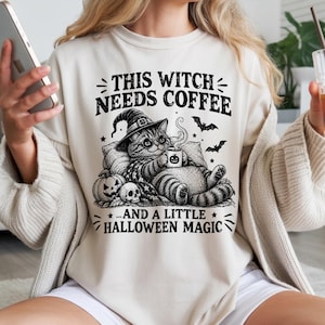 Peut inclure: Un sweat-shirt à col rond couleur crème avec une impression graphique noire représentant un chat portant un chapeau de sorcière, tenant une tasse de café, entouré de citrouilles, d'un crâne et de chauves-souris, avec le texte THIS WITCH NEEDS COFFEE AND A LITTLE HALLOWEEN MAGIC.