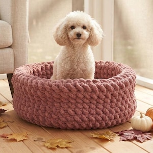 Puede incluir: Un pequeño caniche de color crema sentado dentro de una cama redonda para mascotas de punto grueso en color rosa empolvado. La cama presenta una textura trenzada gruesa y paredes altas y acolchadas, ofreciendo un espacio cómodo para descansar.