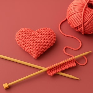 Könnte beinhalten: Ein korallenfarbenes gestricktes Herz, ein Knäuel Wolle und Stricknadeln auf passendem Hintergrund. Die Nadeln halten eine kleine Strickprobe. Die Szene erinnert an handgefertigtes Kunsthandwerk und das Stricken.