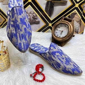 Puede incluir: Un par de zapatillas estampadas en azul y beige con punta. También se ven una pulsera de cuentas rojas y un reloj decorativo. Las zapatillas se exhiben sobre una superficie blanca y esponjosa.