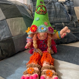 May include: A crocheted gnome doll wearing a green hat with a peace sign, pink glasses, and sandals. The gnome has long pink and blonde hair and is holding a peace sign with its right hand.