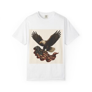 A white short-sleeved t-shirt with a graphic of a bald eagle with outstretched wings carrying a folded American flag in its talons.